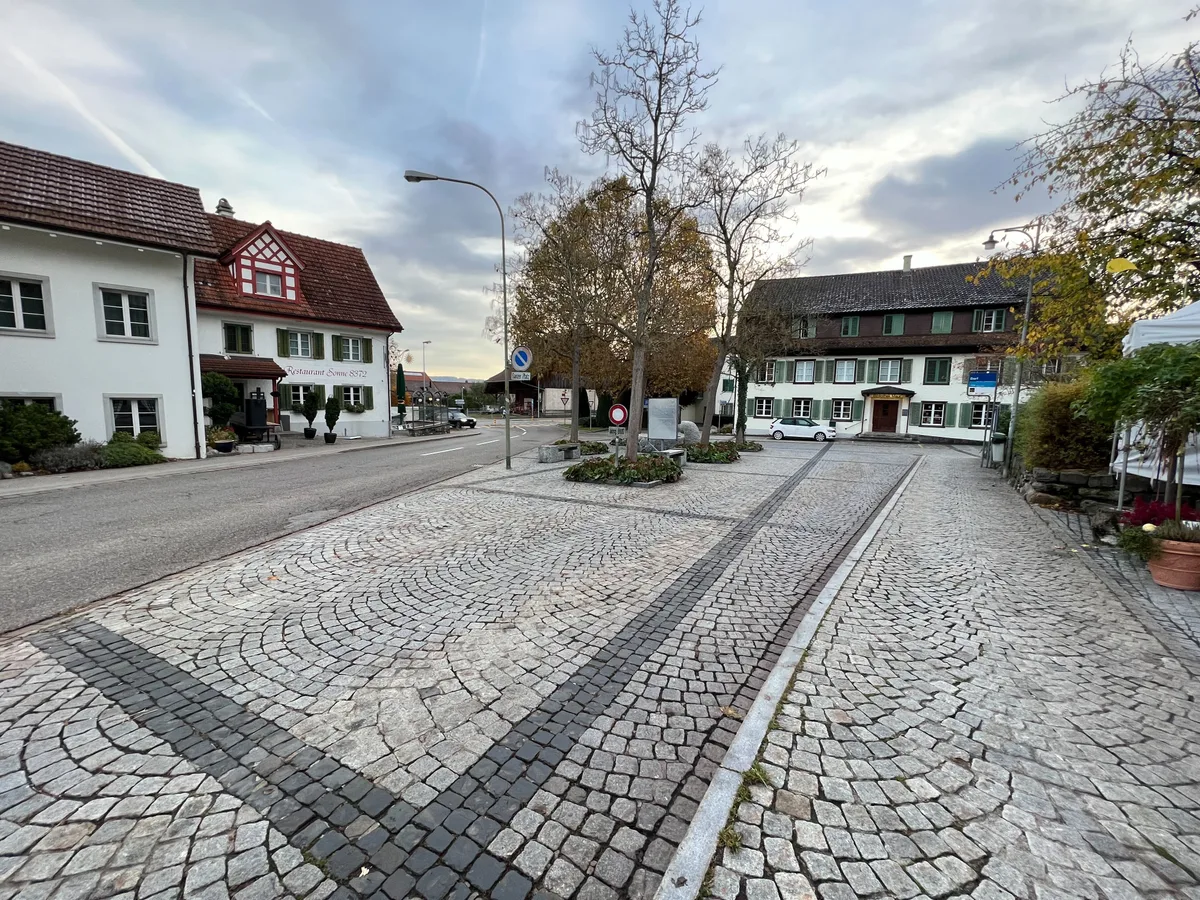 Man sieht eine Strasse durchs Dorf, daneben Häuser, Zebrastreifen, Autos, Busse, Bushaltestellen.